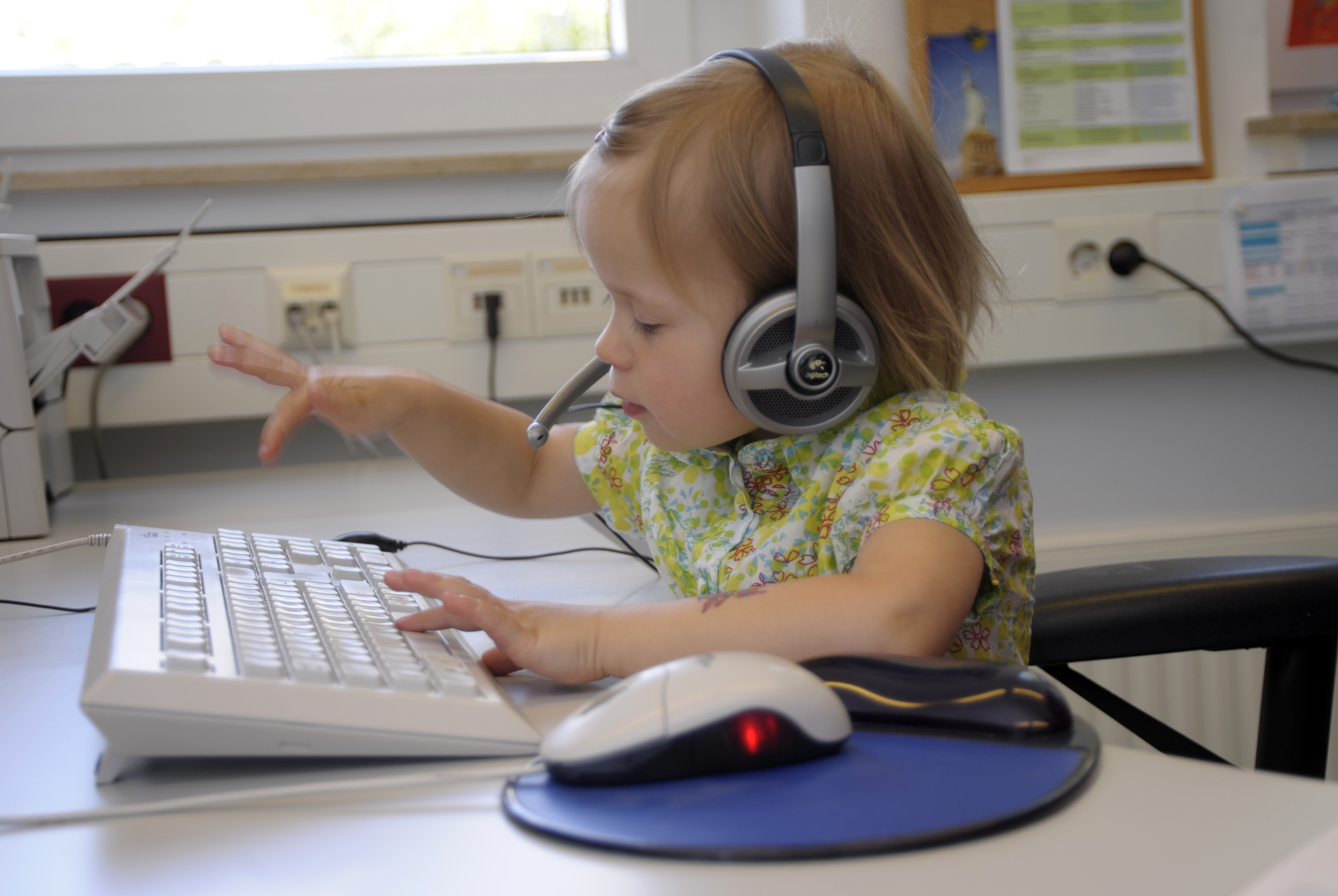 Aufnahme eines blonden Kindes mit einem Logitech-Headset auf dem Kopf, welches an einer weißen Tastatur sitzt. Das Kind ist mitten in der Bewegung des Tippens und wirkt sehr konzentriert.