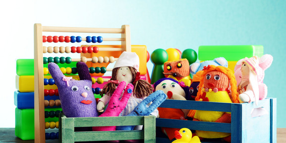 Eine Nahaufnahme verschiedener Spielzeuge in kleinen bunten Holzkisten. Man sieht Stoffpuppen, eine Gummiente, Spielfiguren und einen Abakus mit bunten Holzperlen.