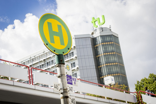 Bushaltestellenschild mit Mensabrücke und Mathetower im Hintergrund.