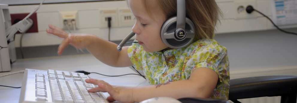 Aufnahme eines blonden Kindes mit einem Logitech-Headset auf dem Kopf, welches an einer weißen Tastatur sitzt. Das Kind ist mitten in der Bewegung des Tippens und wirkt sehr konzentriert.