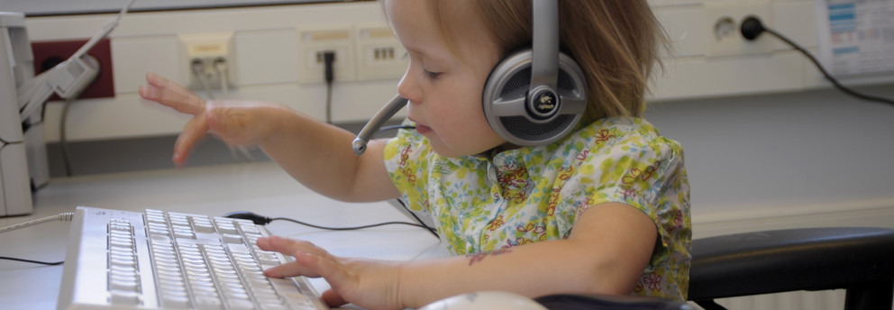 UK-Netzwerk Aufnahme eines blonden Kindes mit einem Logitech-Headset auf dem Kopf, welches an einer weißen Tastatur sitzt. Das Kind ist mitten in der Bewegung des Tippens und wirkt sehr konzentriert.