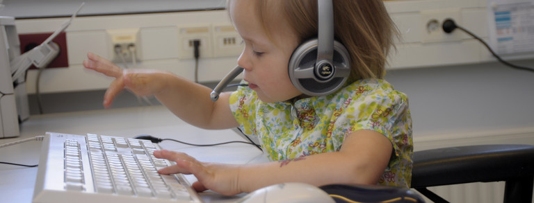 Aufnahme eines blonden Kindes mit einem Logitech-Headset auf dem Kopf, welches an einer weißen Tastatur sitzt. Das Kind ist mitten in der Bewegung des Tippens und wirkt sehr konzentriert.
