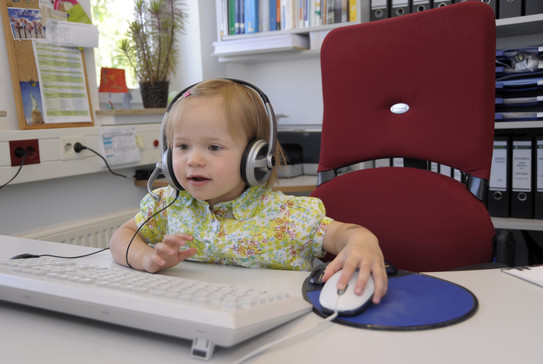 Kleinkind mit Kopfhörern bedient Maus am PC Arbeitsplatz.